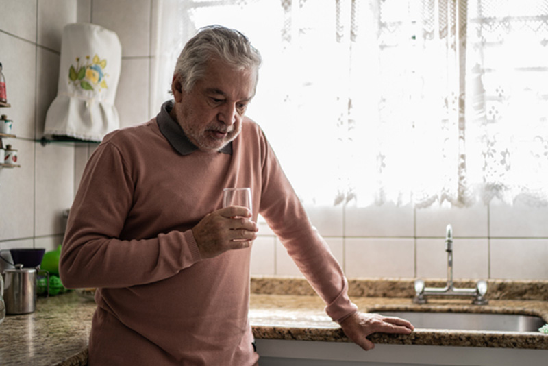 A depressed man stands in the kitchen with a glass of water, an important reminder of the association between depression and heart health.